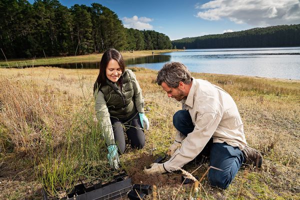 A young woman and man, wearing warm outdoor clothing are kneeling in the grass on the edge of a reservoir, caring for the land.